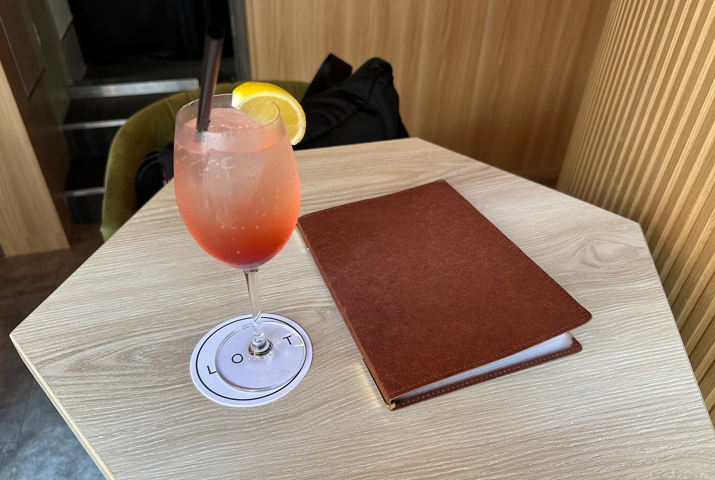 En ljusröd drink står på ett ljust hexagonalt bord där det också ligger en brun drinkbok.