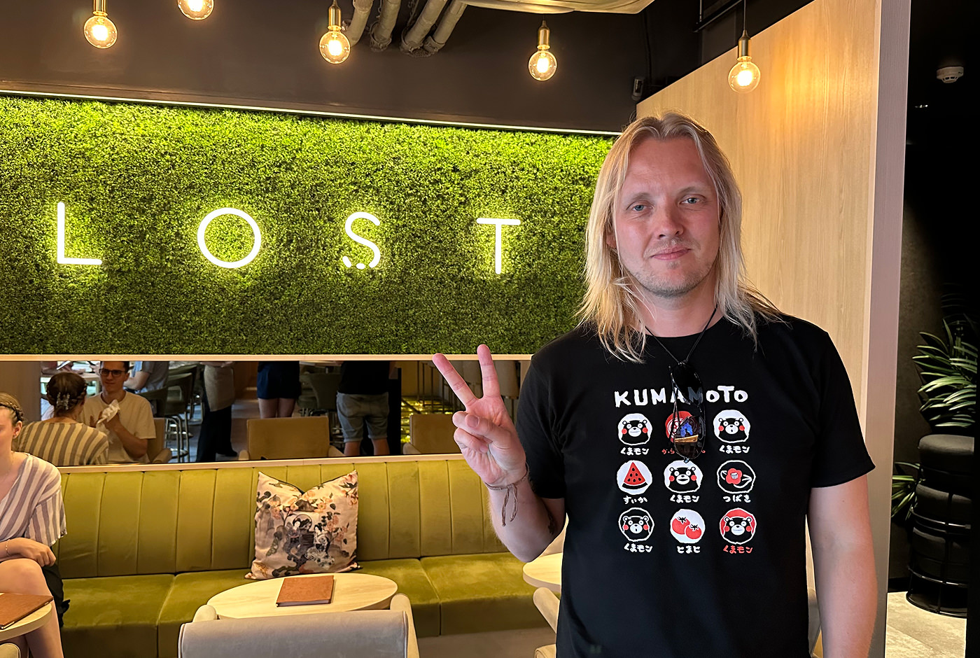 Jag står iförd en svart t-shirt och ger fredstecknet åt kameran. Jag står i en upplyst bar med en gräsvägg med en skylt gjord av ljusa slingor med texten "Lost".