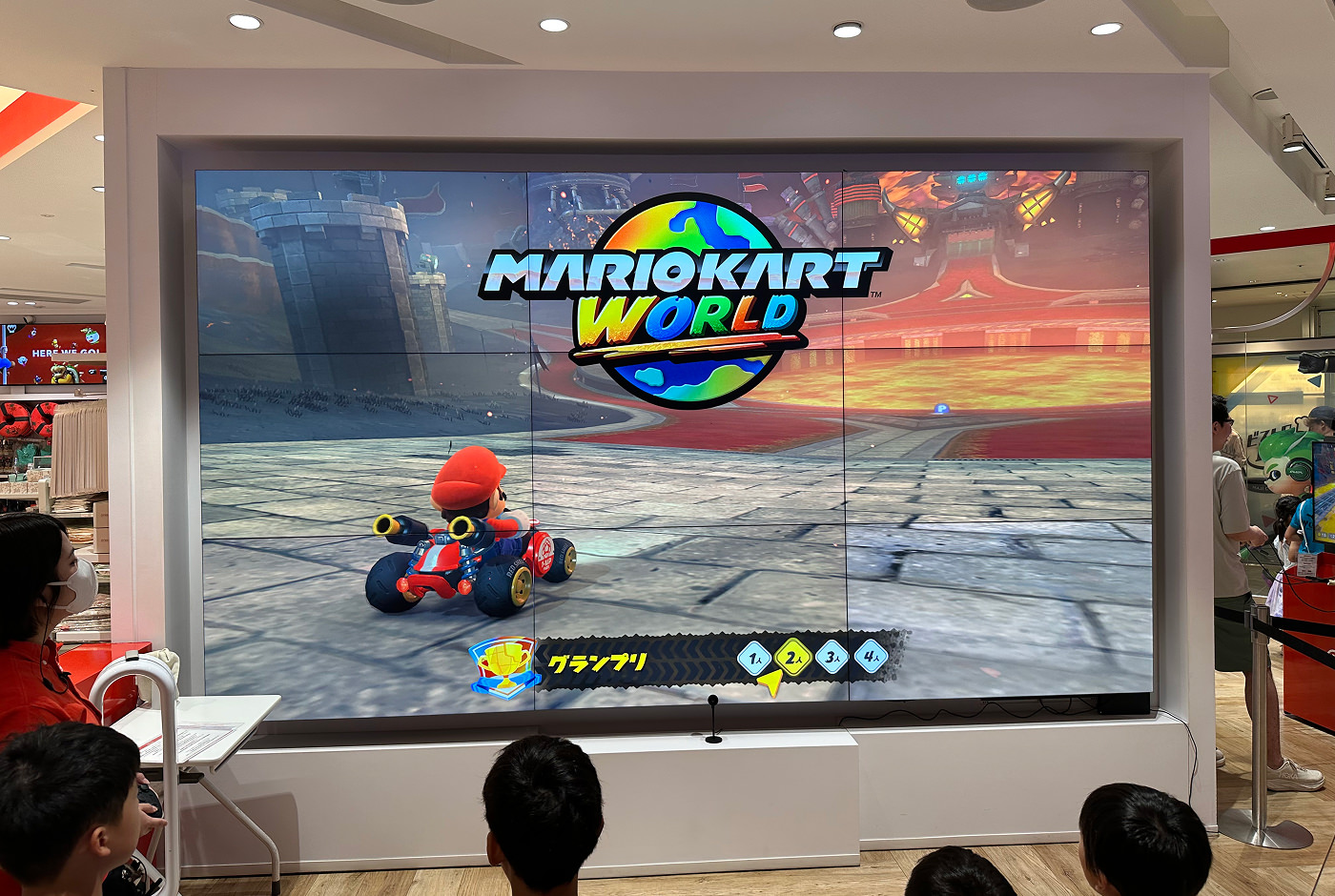 En enorm TV-skärm i en butik där TV-spelet Mario Kart World är igång. Man ser Super Mario sitta i en gokart och köra på en väg.