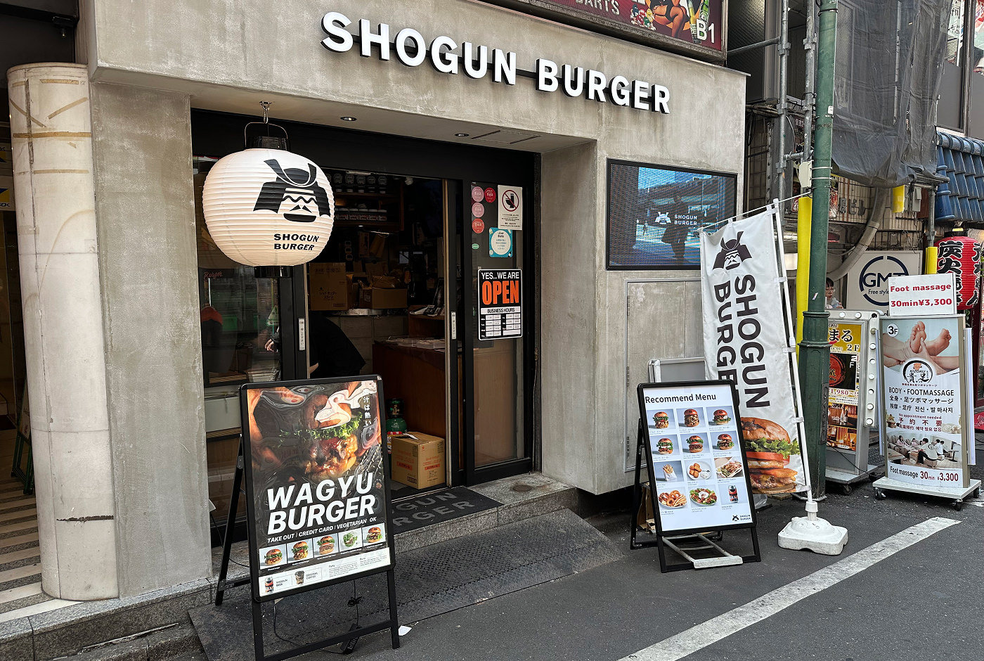Grå fasad till en liten restauran med glasdörr. Utanför står det "Shogun burger" på flera skyltar och affischer.