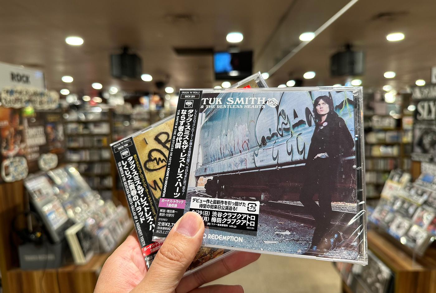Hand håller i två CD-skivor inuti en stor skivbutik. Ena skivan har en bild på en mörkt klädd man som står vid en tågvagn. Titeln lyder "Tuk Smith & The Restless Hearts".