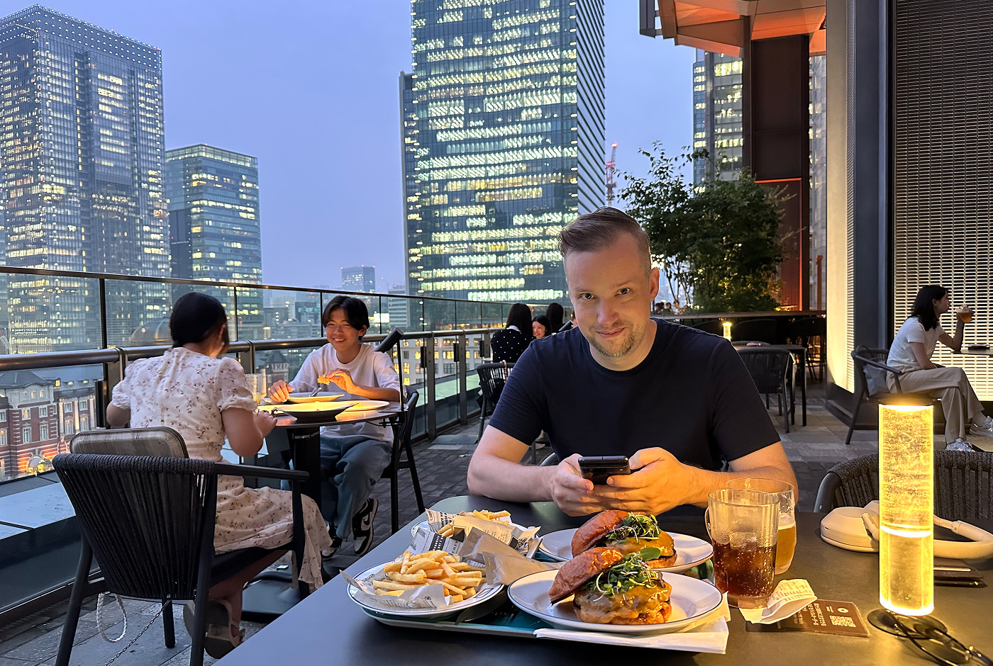 Dan sitter på en uteservering i mörk skjorta och ler finurligt åt kameran. På bordet står två portioner med hamburgare och pommes frites. I bakgrunden syns upplysta skyskrapor.