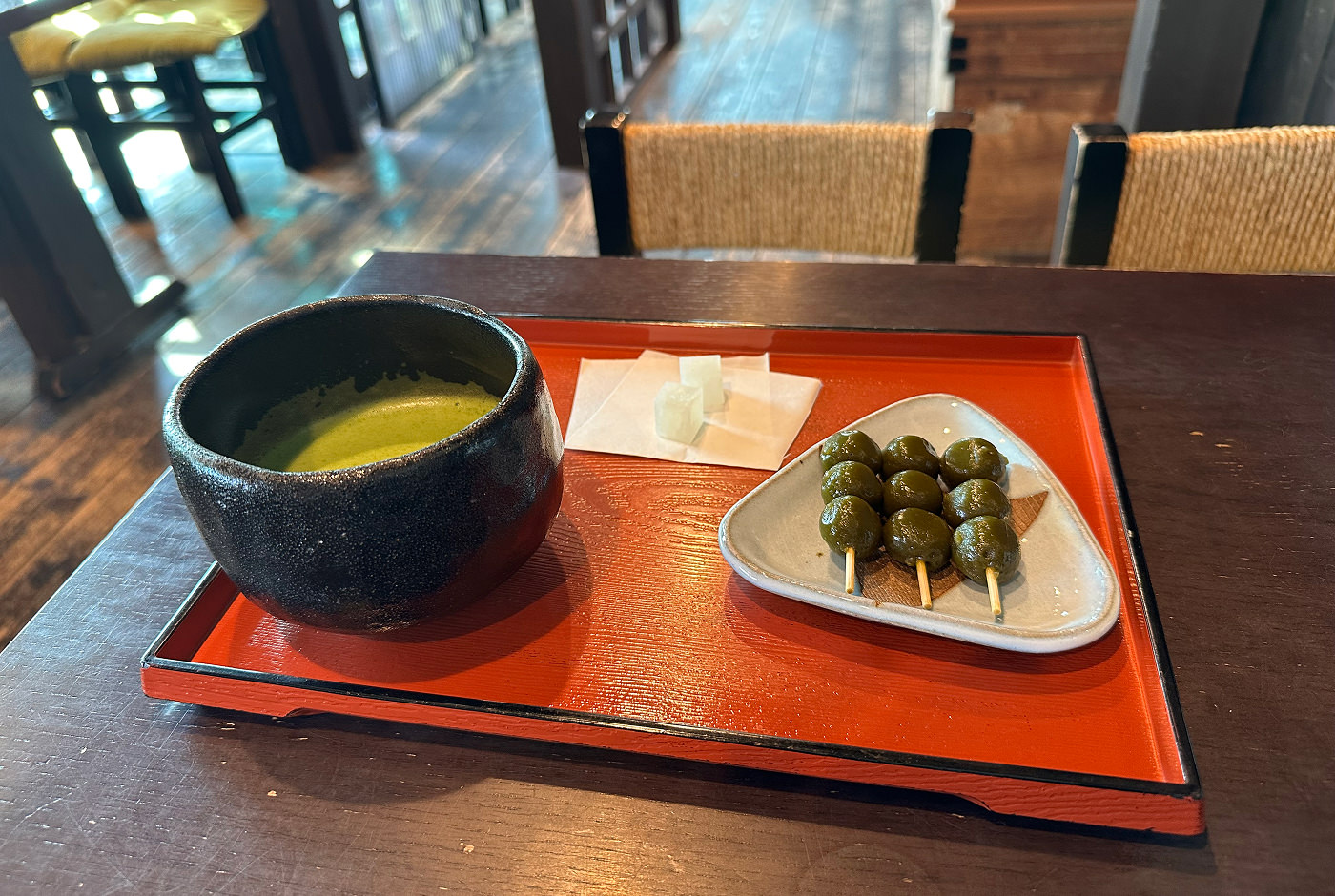 Röd träbricka med en svart kopp med grönt matche-te och en vit tallrik med tre pinnar med tre gröna matcha-dumplings på.
