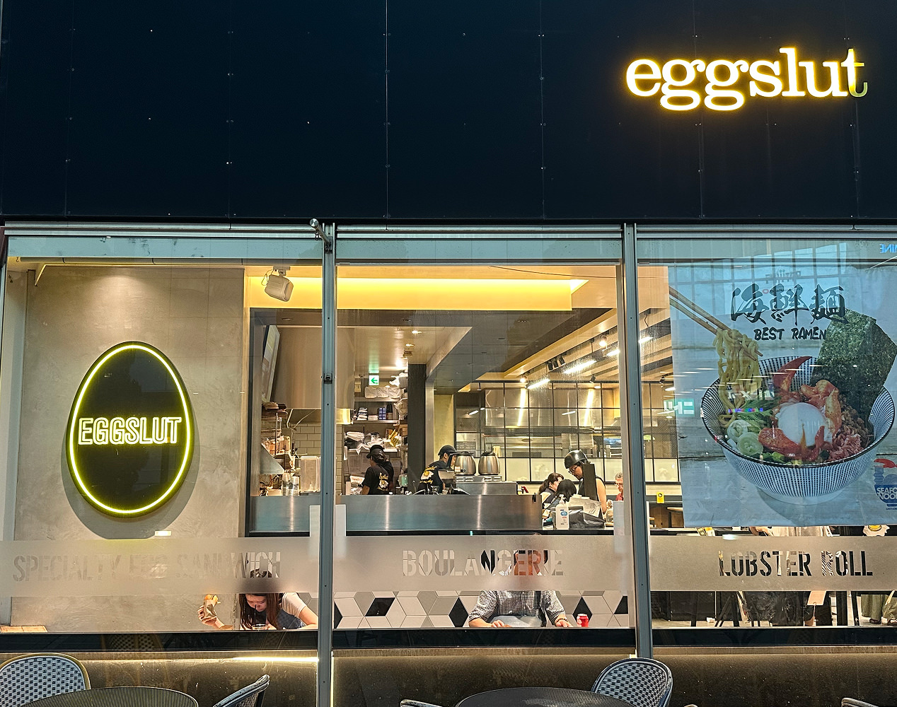 Fasad till en restaurang som främst består av fönster. Två skyltar har gul text och svart bakgrund och lyder "Eggslut".