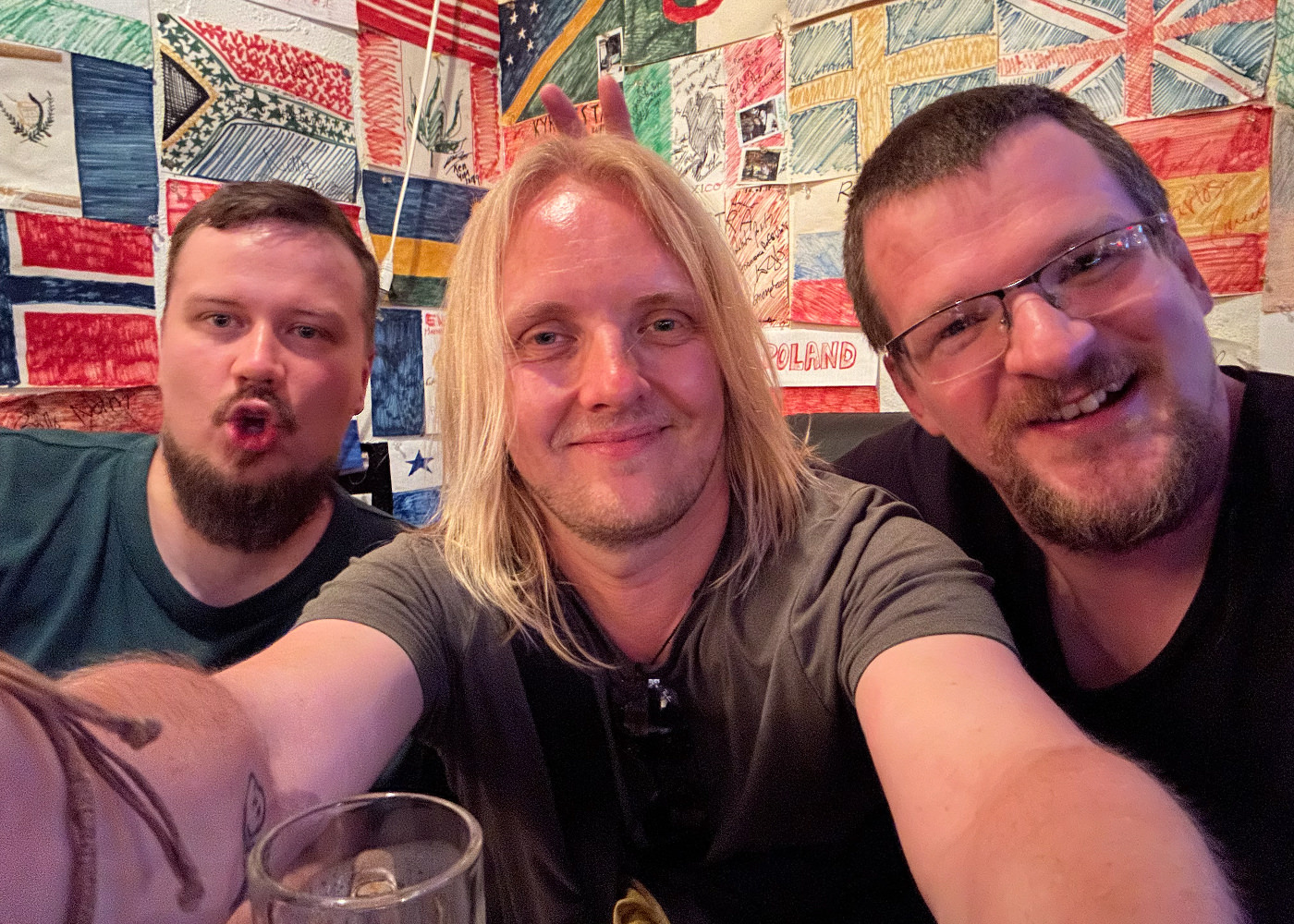 Jag ler ordentligt och tar en selfie med två andra män som båda har kort brunt hår. han ena har glasögon. På väggen bakom oss hänger ritade flaggor.