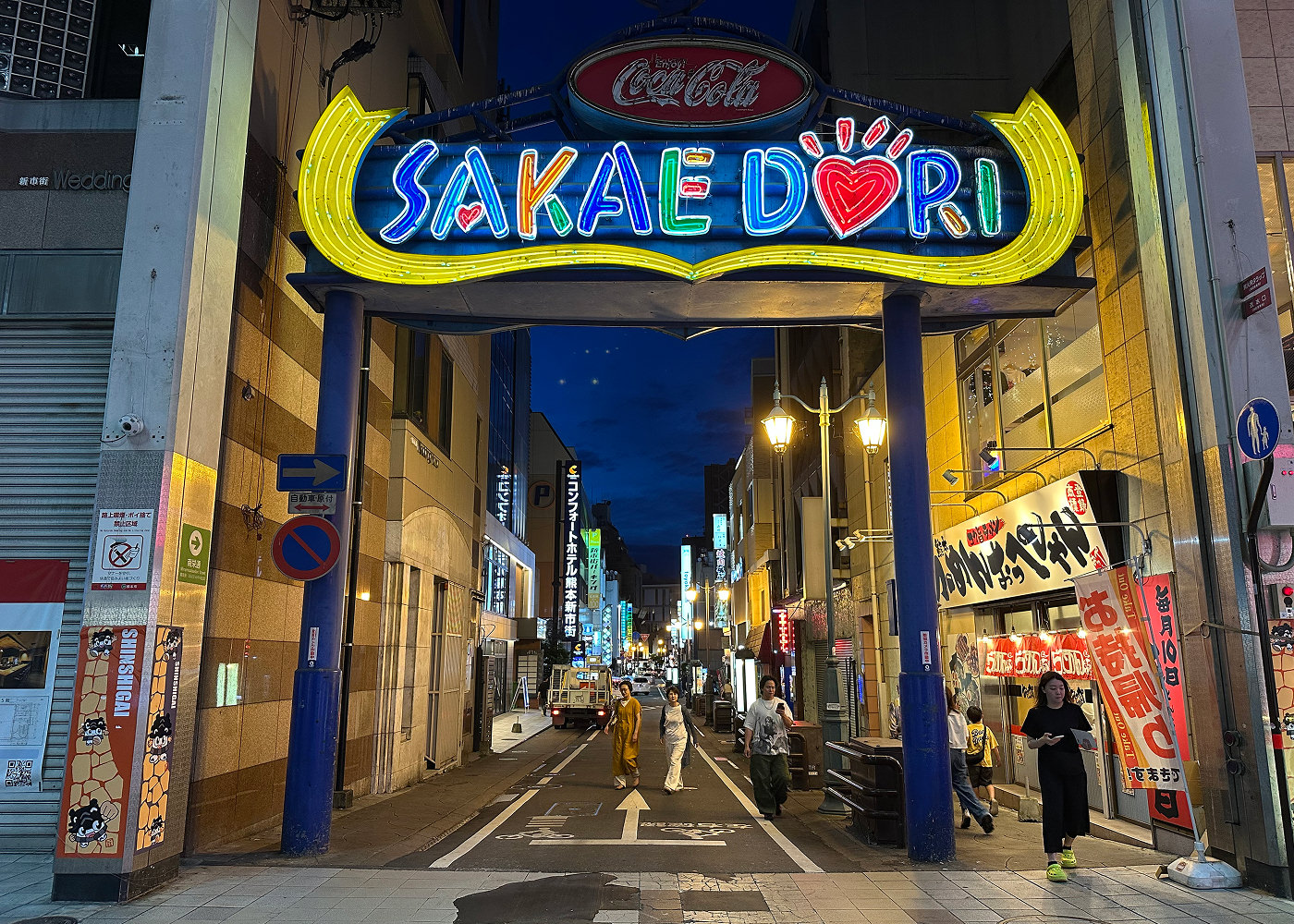 Ingång till en gågata med matställen och barer på båda sidorna. Ingen består av en blå port med en neonskylt i olika färger med texten "Sakae Dori".