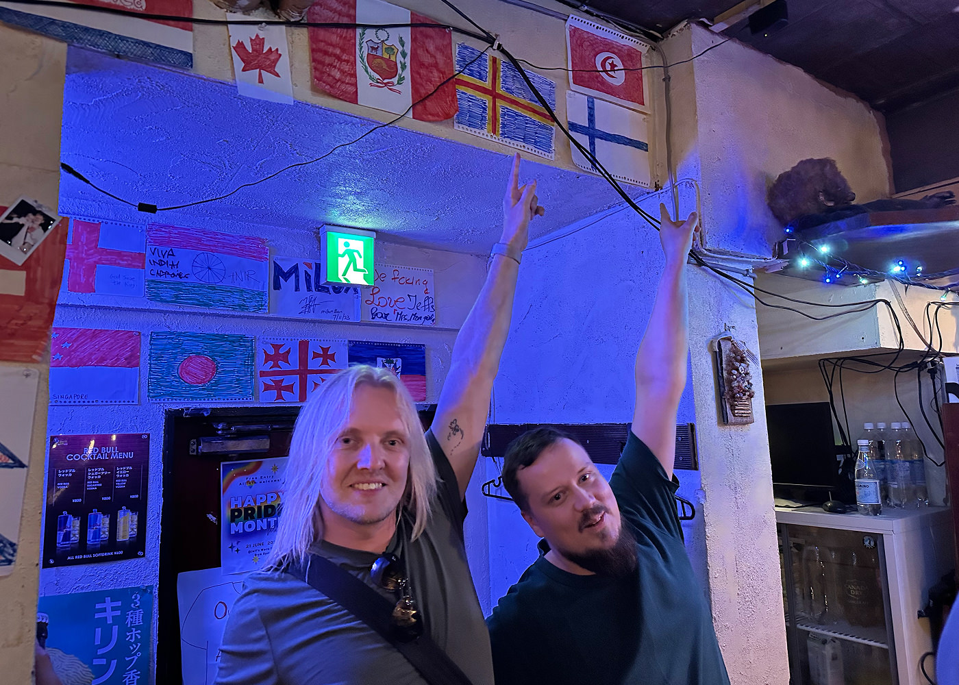 Jag och en annan västerlänning står och pekar upp på en bars vita vägg. På väggen hänger flera ritade flaggor. Vi pekar på Finlands och Ålands flaggor.