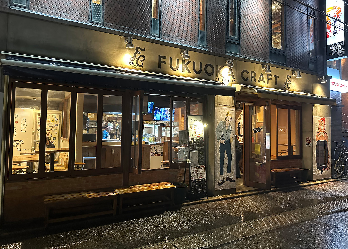 Upplyst fasad med stora fönster. Skylt ovanför ingången lyder "Fukuoka Craft". Det är mörkt ute och gatan är blöt.