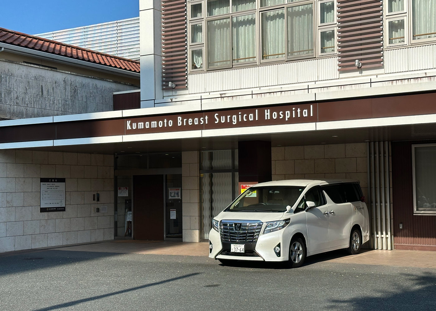 Infarten till ett sjukhus med en skylt som lyder "Kumamoto Brest Surgical Hospital". En bil står parkerad utanför.
