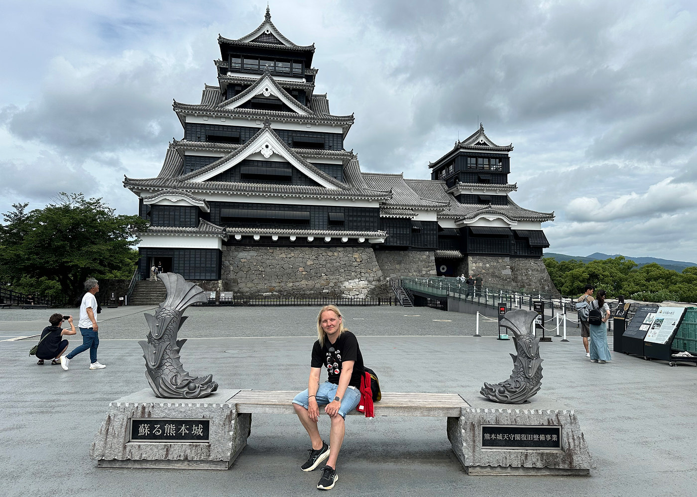 Jag sitter på en träbank på ett öppet torg framför ett svart och vitt japanskt slott med ett stort och ett litet torn.