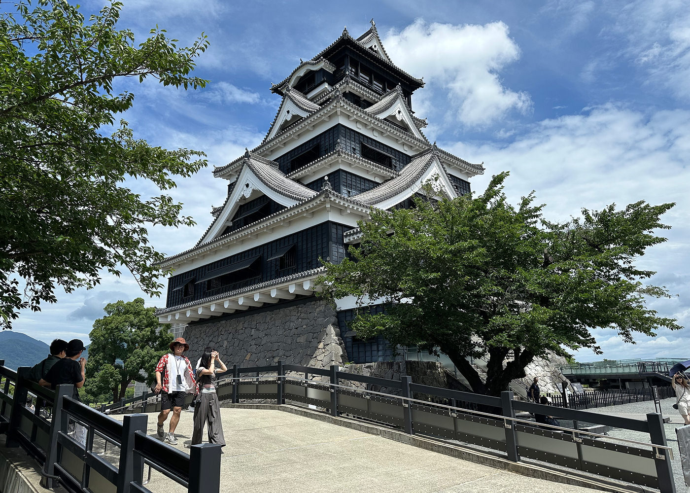 Svart och vitt japanskt slottstorn mot lätt molnig himmel. I förgrunden går fyra personer på en liten bro.