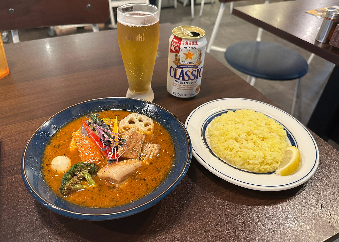 En mörk skål med orange currysoppa med grönsaker står på ett bord bredvid en vit tallrik med ris och ett upphällt ölglas.