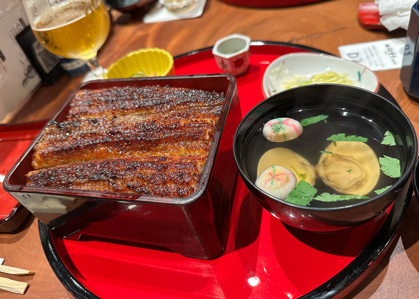 Gyllenbrun grillad ål i en mörk låda. Till höger står en svart skol med genomskinlig musselsoppa.