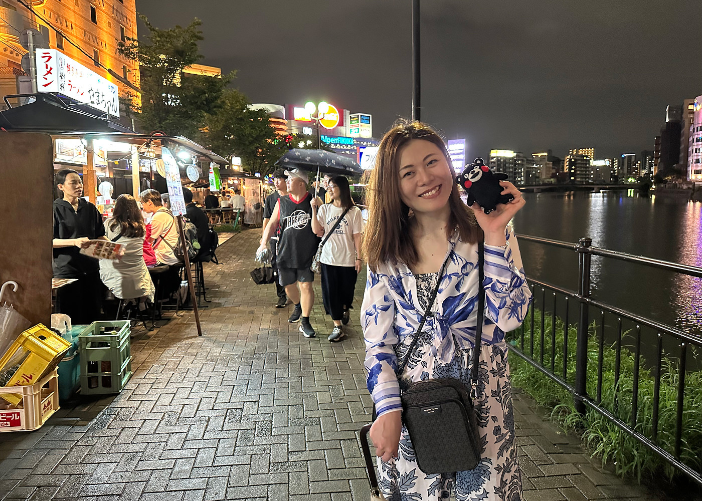 Vacker japansk kvinna i ljus klänning ler in i kameran. Hon står på en gågata med matstånd längd med en flod.
