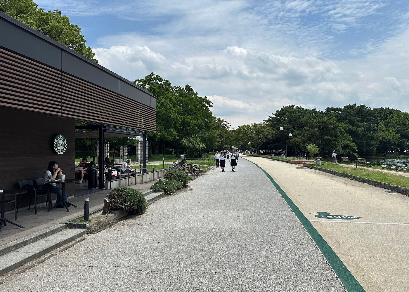 Lång promenadgång i sten genom en lummig park. Till höger skymtas vatten. Till vänster sitter en tjej på en stol vid ett Starbucks-fik.