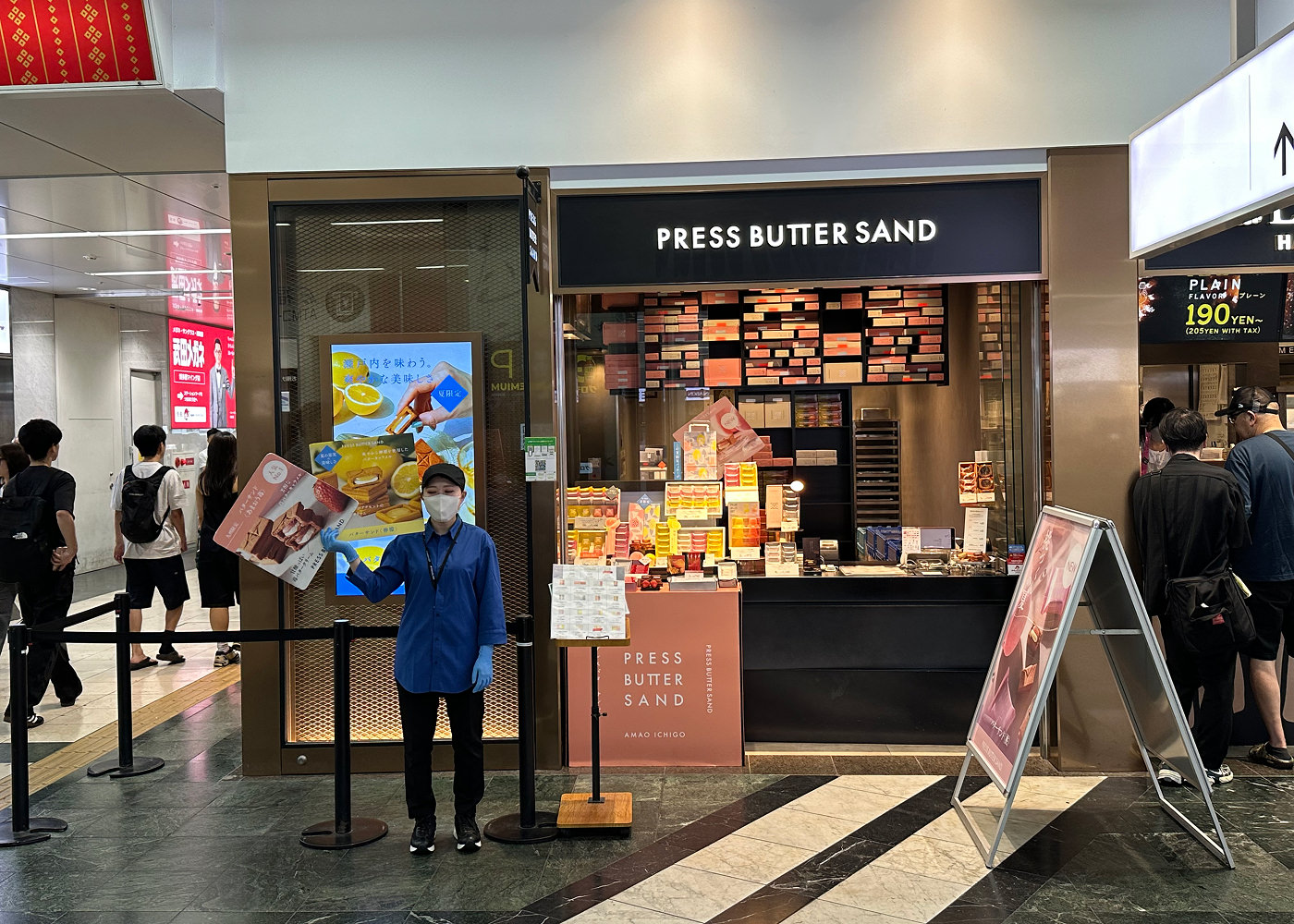 En liten butik i en tågstation med hyllor med många papplådor. En skylt ovanför lyder "Press Butter Sand".