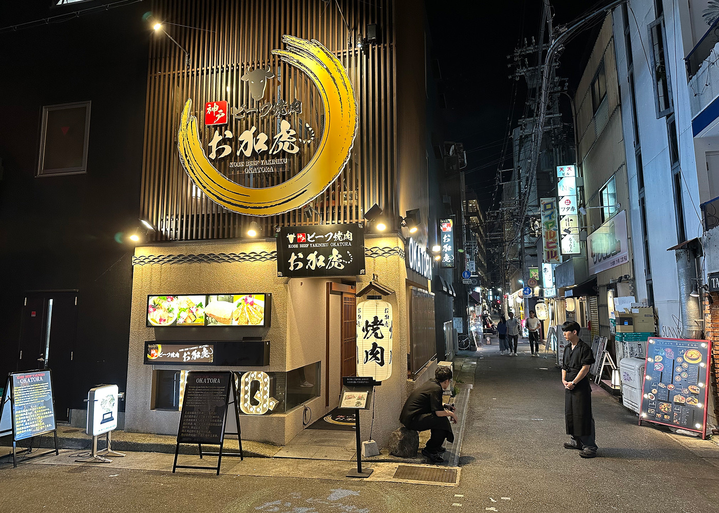 Två svartklädda servitörer står vid en upplyst gatukorsning utanför en restaurang med en stor skylt som består av ett gultrunt penseldrag och texten "Kobe Beef Yakiniku Okatora"inuti. Det är mörkt ute.