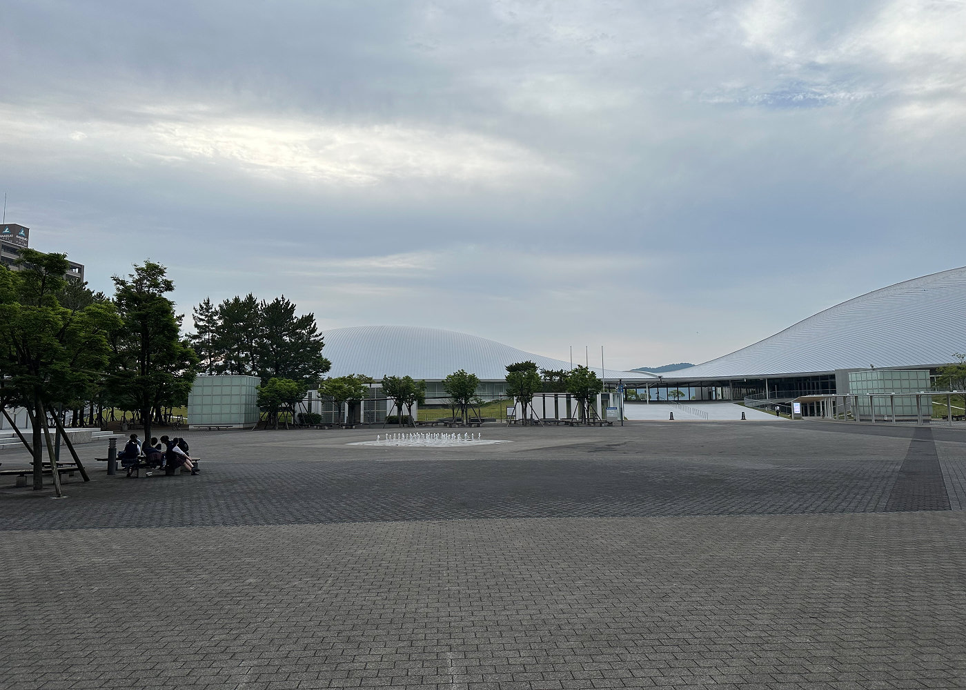 Stort öppet stenbelagt torg. Längst bort syns en rund inomhusarena och till vänster står flera träd planterade i utkanten av torget.