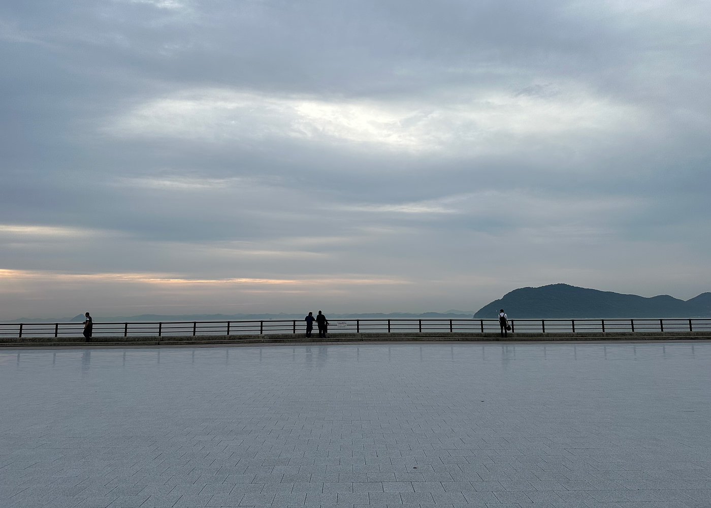 Stor och bred stenpromenad vid ett hav. Fyra personer står vid räcket vid havet. I fjärran skymtar lummiga stora öar.