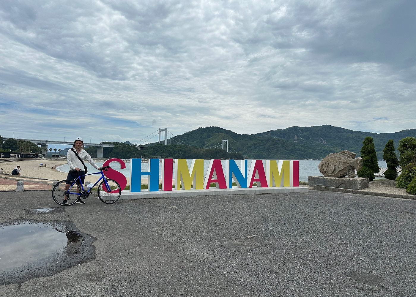 Jag står och håller i en cykel framför en skylt framför en sandstrand. Skylten lyder "Shimanami". I fjärran ser man en hängbro och lummiga berg.