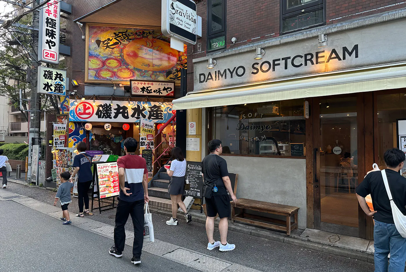 Sju personer går och stor vid en gata med två butiker. En fasad är färggrann med japansk skrift och en annan ljusgrå med texten "Daimyo Softcream".