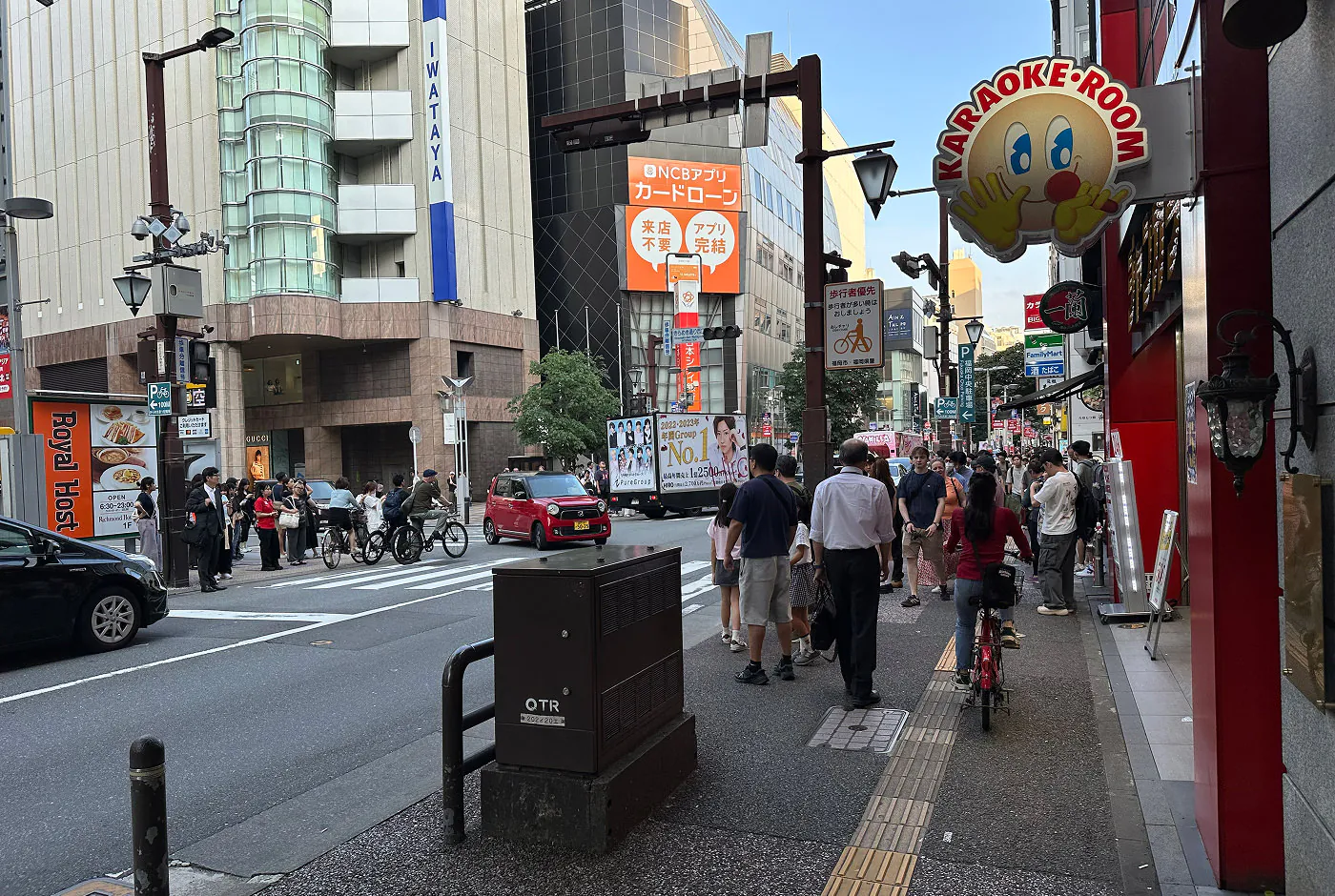 Korsning i en upptagen del av en stadskärna. Trottoarerna är fulla med folk och bilar kör på gatan. En skylt i förgrunden består av ett leende ansikte och texten "Karaoke Room".