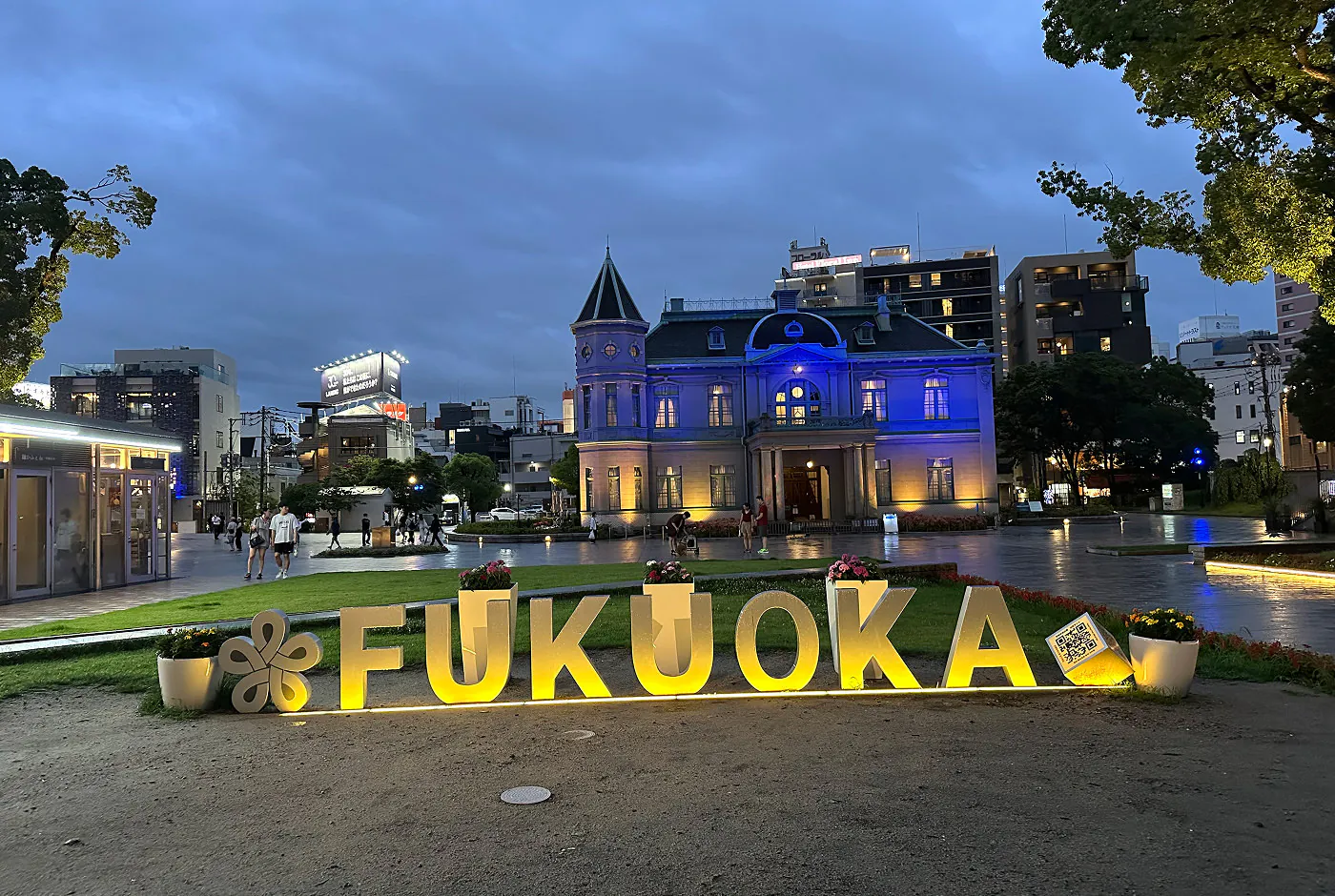 En upplyst gul skylt på marken som lyder "Fukuoka". Bakom den finns ett öppet torg och ett gammalt medborgarhus.