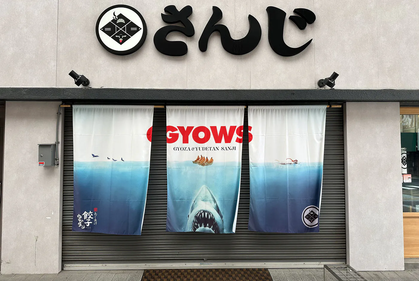 Stängd bred ingång till en restaurang. Tygstycken hänger framför ingången med bild på en haj och texten "Gyows".