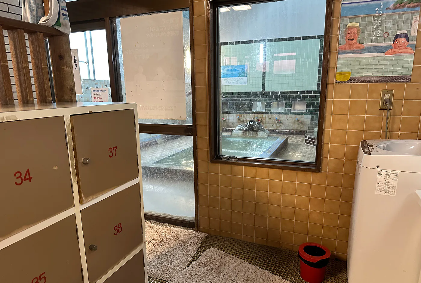 Omklädningsrum med skåp och tvättmaskin i ett litet badhus. Via ett fönster kan man se badet som har en rektangulär pool i mitten.