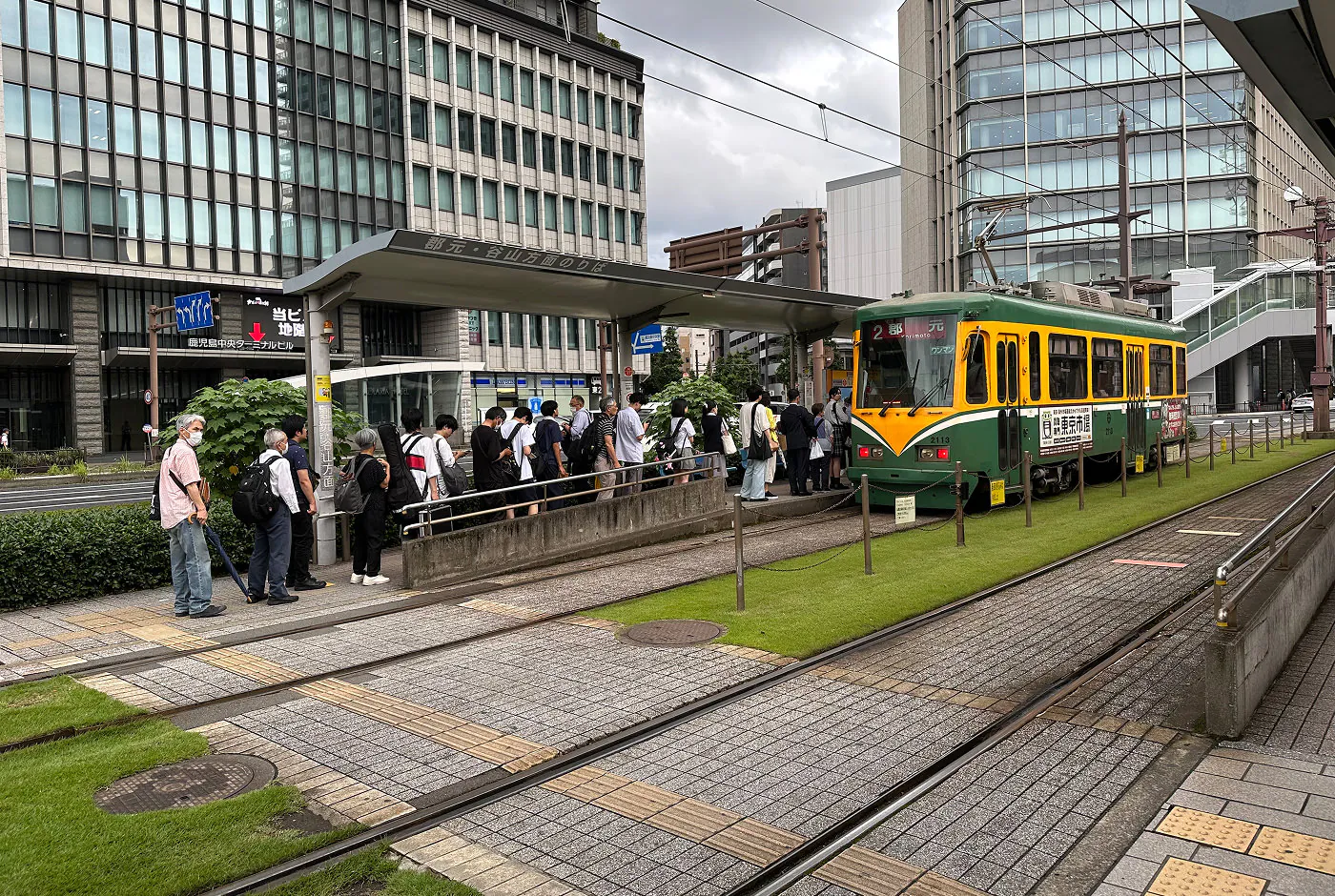 Cirka 20 personer köar till en grön och gul spårvagn. Marken runtom består av välansat gräs och stenplattor. I bakgrunden finns höghus.