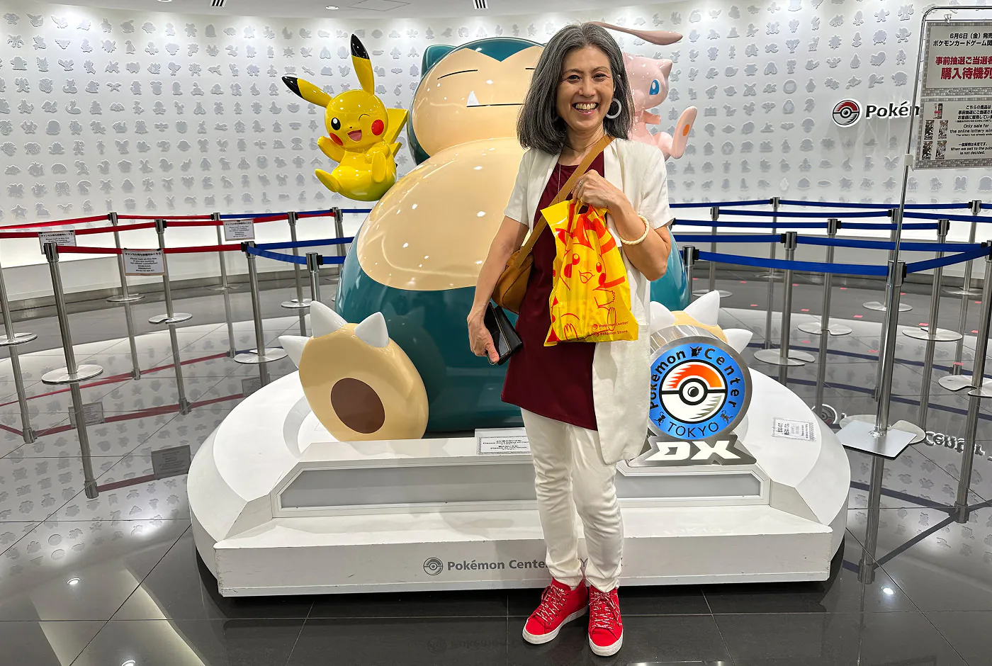 Keiko håller i en gul påse och står framför en stor staty över tre pokomons. En skylt lyder "Pokemon Center Tokyo".