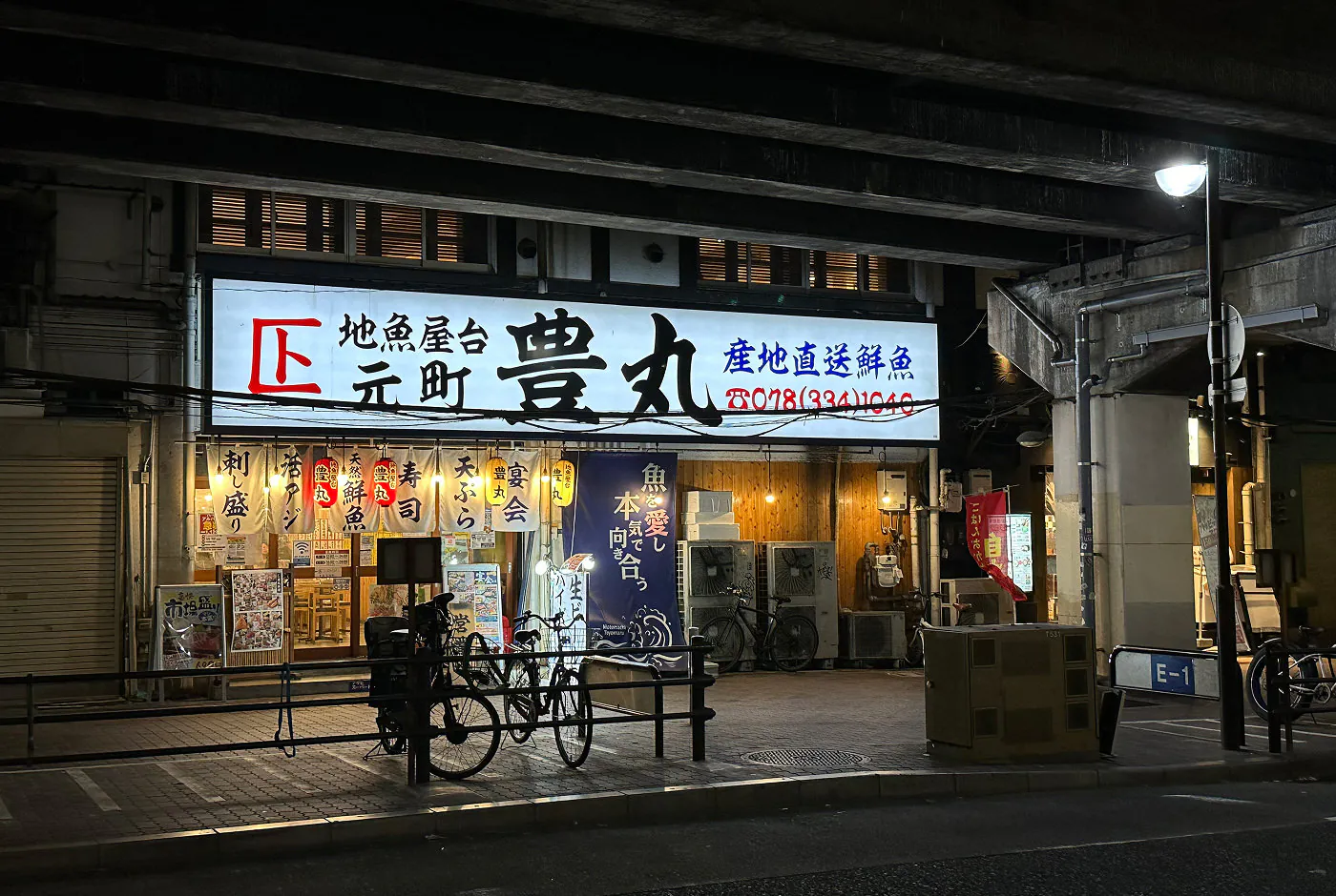 Ingång till restaurang under en tågstation. Ingången är upplyst med en bred vit skylt och flera vita dukar. Alla har japansk skrift. Framför är två cyklar parkerade