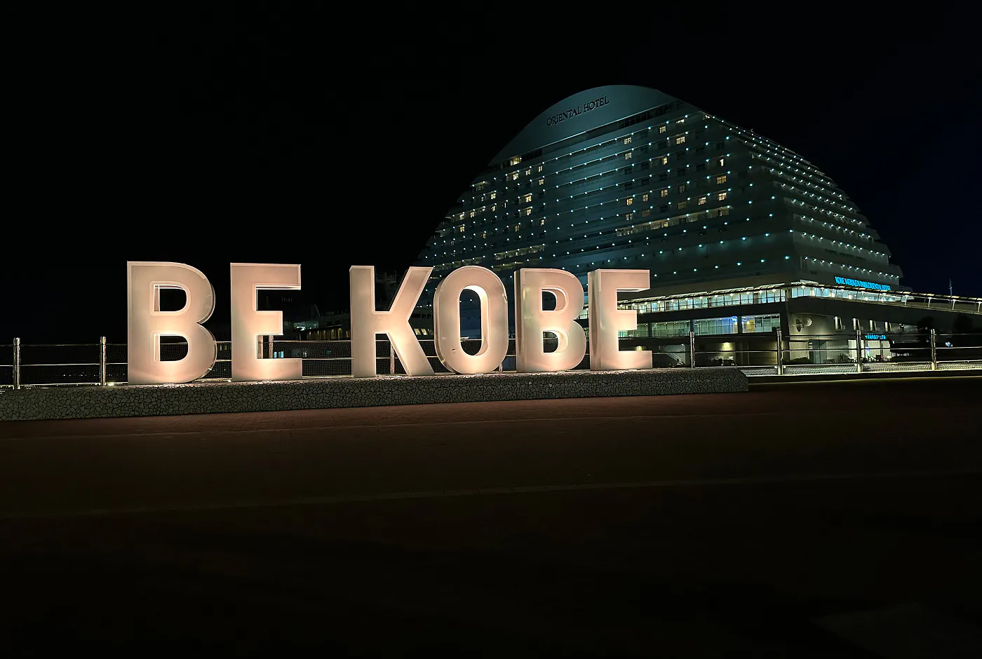 Fyra stora bokstäver som bildar texten ”Be Kobe”. Bokstäverna är upplysta. I bakgrunden står ett stort och upplyst ovalt hotell.