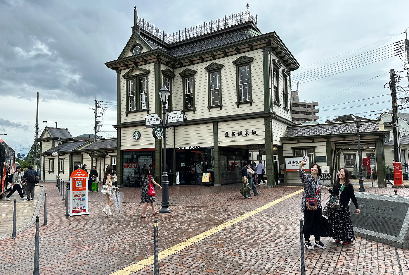 En gammaldags tågstation med europeisk arkitektur. Byggnaden är ljus ed gröna knutar. Två kvinnor skrattar ochb tar en selfie vid sidan.