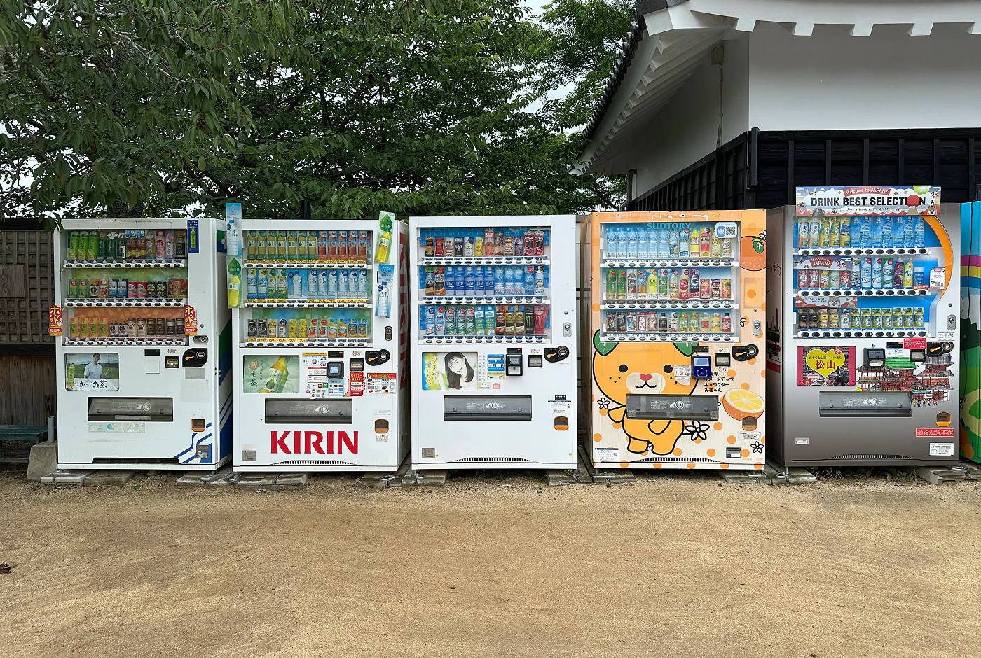 Fyra dryckesautomater jämte varandra på en grusplätt. Bakom växer ett buskigt träd.