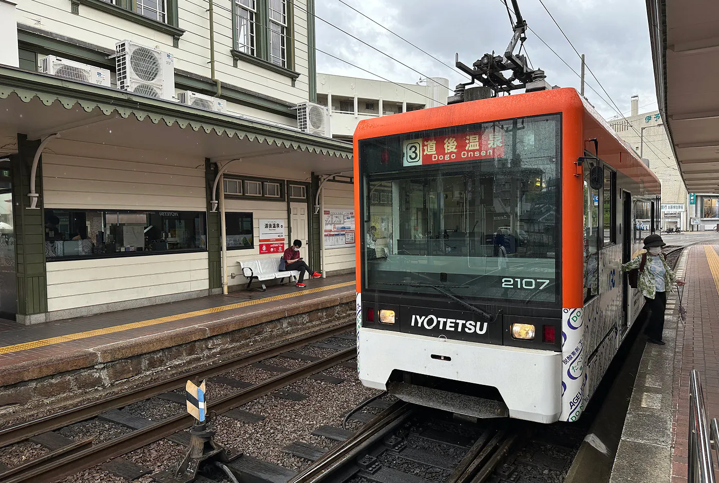 En vit och orange spårvagn vid en station mellan vita hus. Skylten lyder "3 Dogo Onsen".