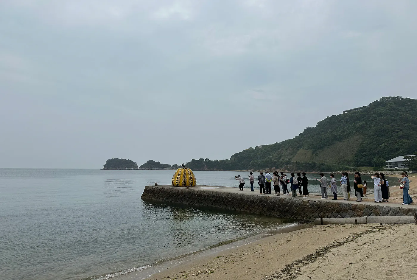 Flera japaner står i kö på en pir vid en strand för att fotografera en stor skulptur av en gul pumpa med svarta prickar.