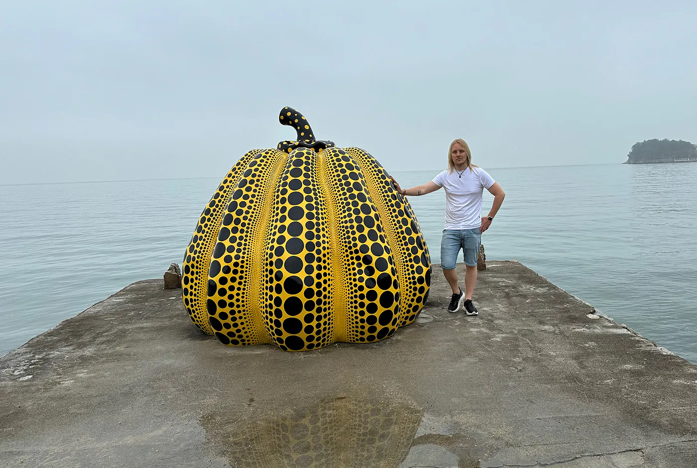 Skulptur av en stor gul pumpa med tusentals svarta prickar. Den står på en betongpir framför ett dimmigt hav. Jag står bredvid den och håller handen mot den.