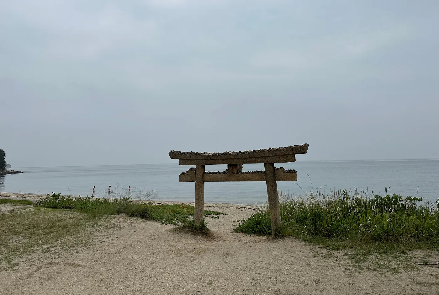 Gråbrun torii-port nere vid en sandstrand. I vattnet badar tre personer.