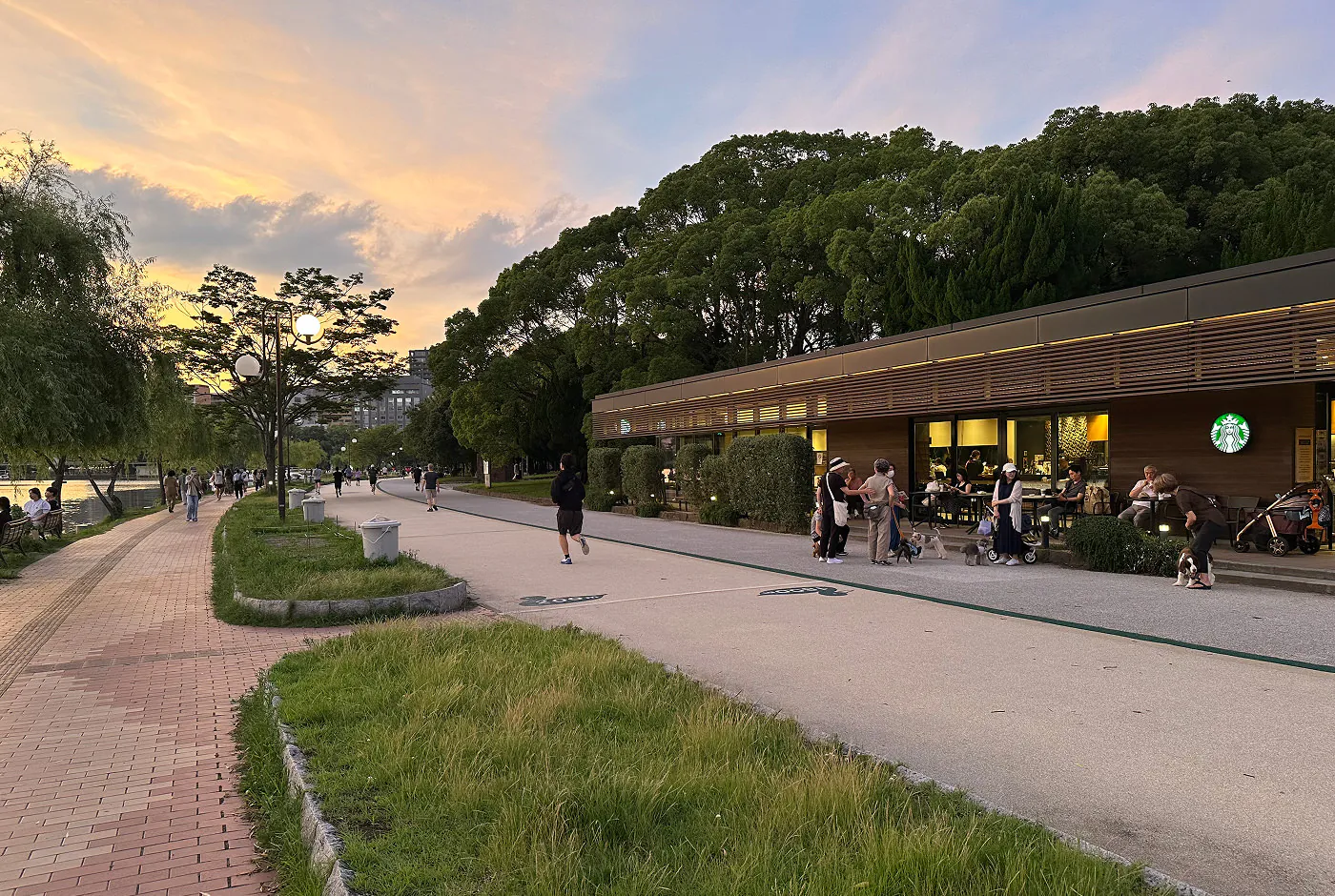 Park med två långa promenadgångar separerade av ett parti med gräs och träd. Besökare joggar och promenerar. Vissa sitter vi en lång brun byggnad med logotypen för Starbucks på.