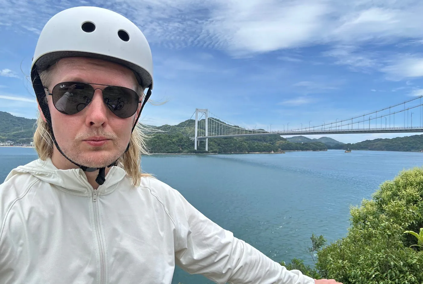 Jag har vit jacka, vit cykelhjälm, solglasögon och tar en selfie. Bakom mig är vit bro mellan två öar i ett hav.