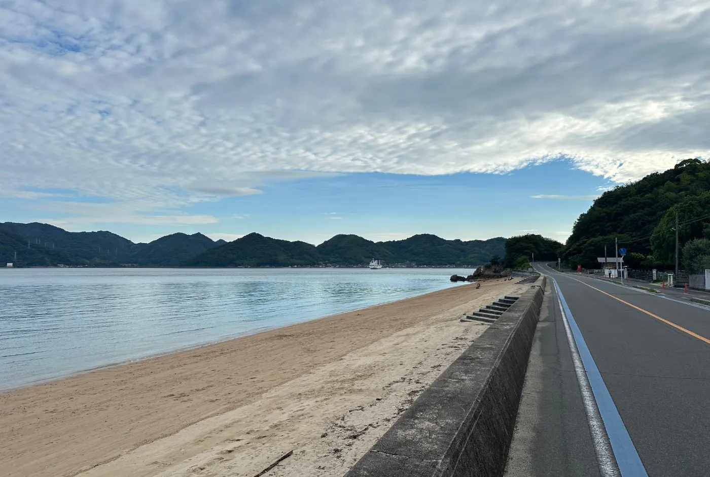 Bil- och cykelväg längs med en sandstrand. På andra sidan finns en lummig bergskedja.