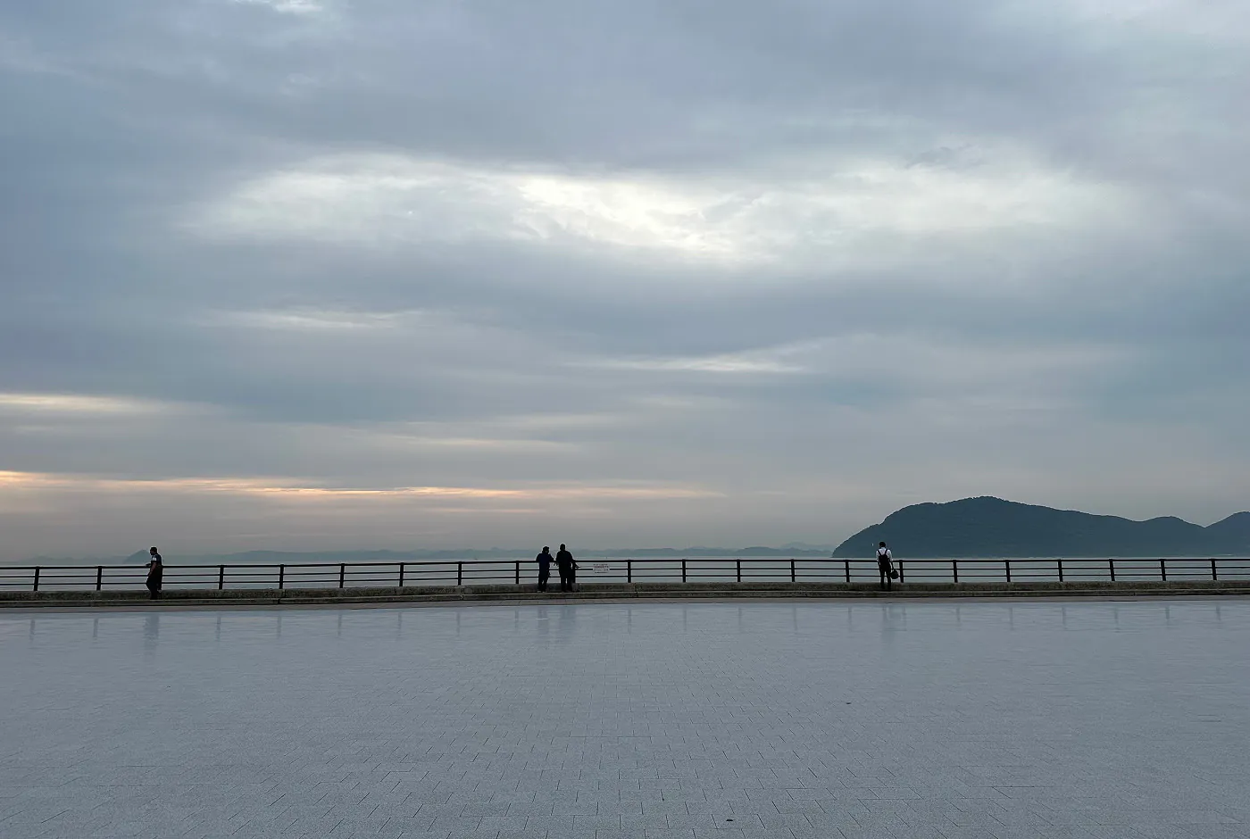 En riktigt stor öppen yta vid en stenpromenad. Himlen är dimmig och i fjärran syns lummiga berg. Fyra personer står vid ändan.