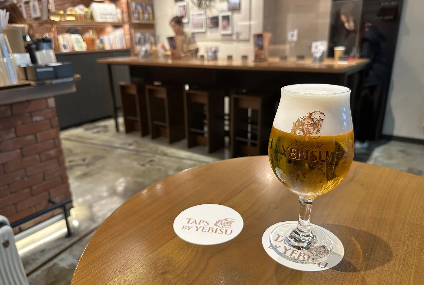 Ett runt glas öl står på ett runt träbord. Ett vitt drinkunderlägg har texten "Taps by Yebisu".