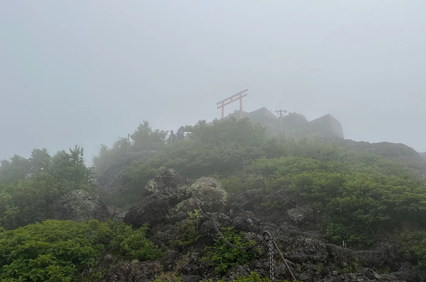 Lummigt och stenigt berg med en röd torii-port på toppen jämte ett hus.