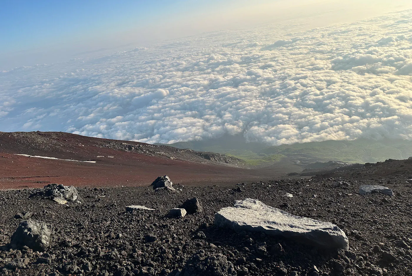 Massiv sluttning bestående av grå och röd vulkansten ned till en lummig dal. I fjärran syns moln ovanifrån.