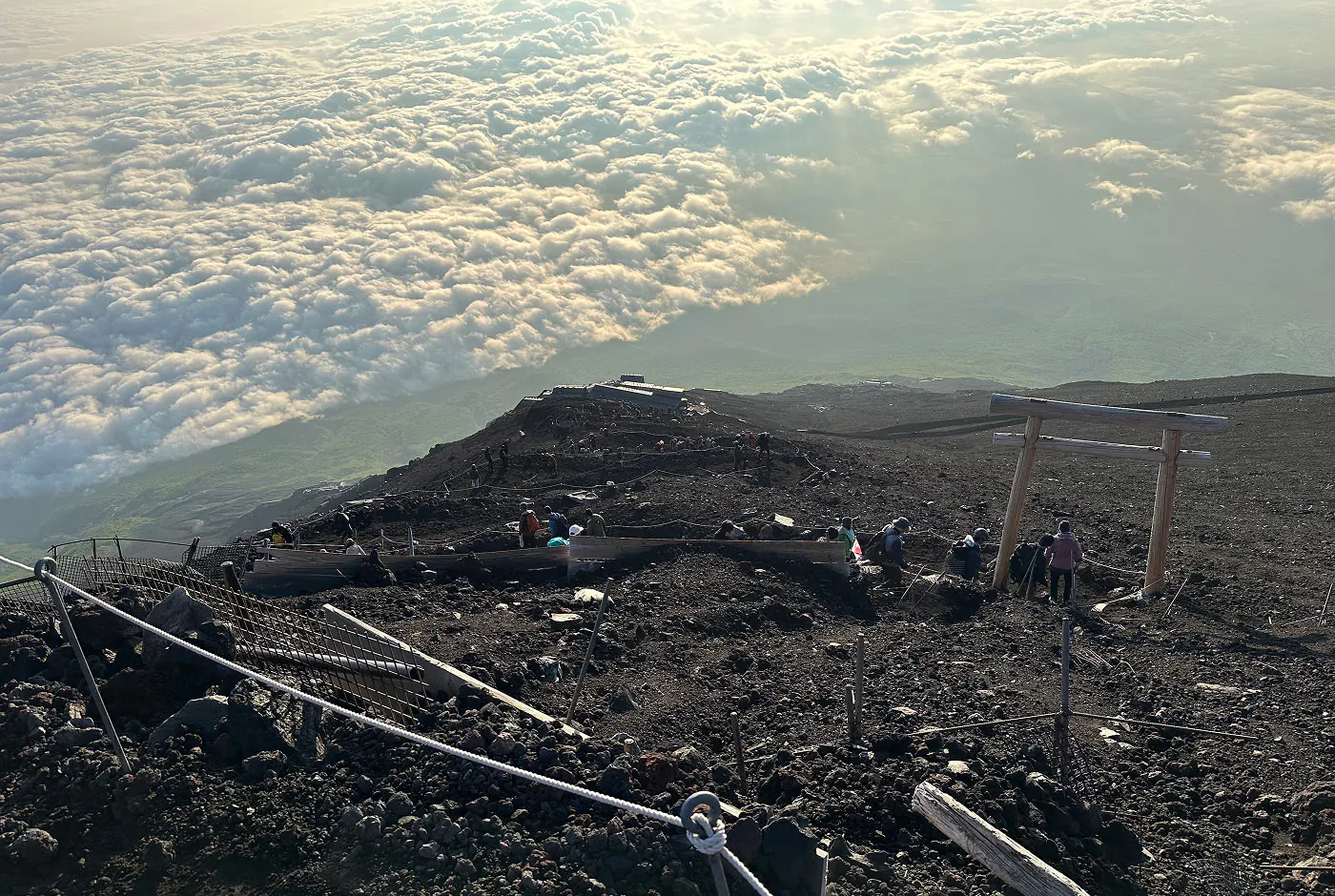 Vandringsled upp för ett berg som går i sicksack.Folk promenerar upp. I fjärran syns en lummig dal och moln.
