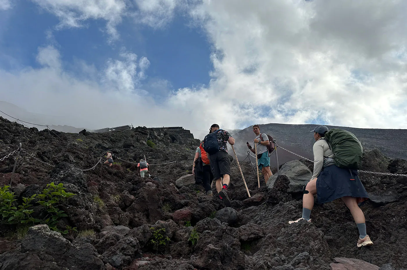 Väldigt brant och stenig backe upp till ett hus på ett berg. Sju personer håller på att går upp med vandringsstavar.
