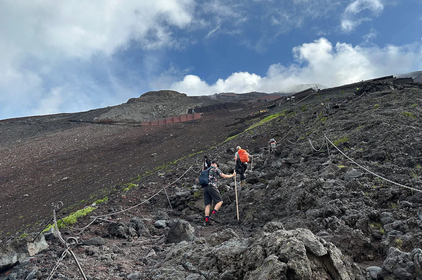 Brant och stenig vandringsled rakt upp för ett enormt berg. Tre personer håller på att går upp för leden. I toppen finns det byggnader.