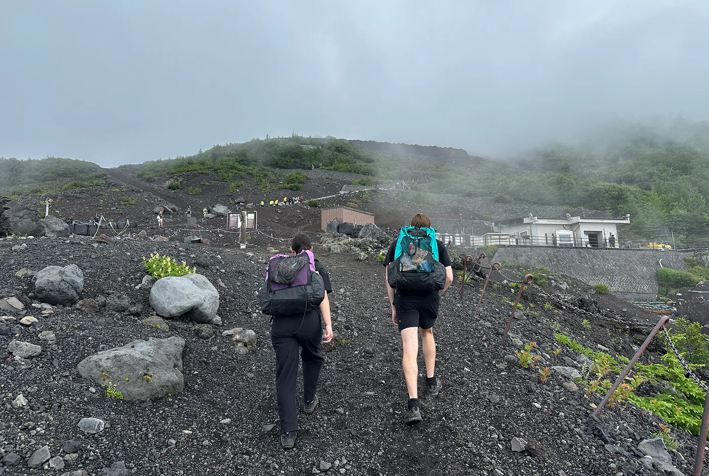 Vandringsled går uppåt över kulle med grå vulkansand och em del gröna buskar. Ett par med varsin ryggsäck och svarta kläder promenerar uppåt.